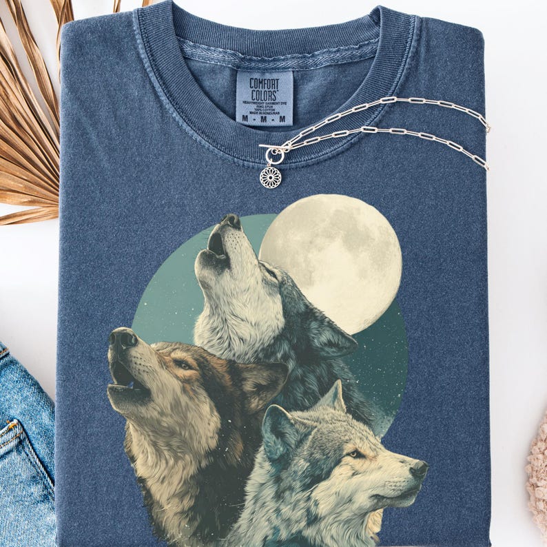 May include: A blue Comfort Colors t-shirt featuring a graphic of three wolves howling at a full moon. The shirt has a crew neck and short sleeves. A silver necklace with a floral pendant is draped over the shirt.