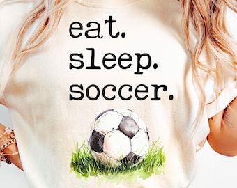 May include: Cream-colored t-shirt with the words "eat. sleep. soccer." in black. Below the text is a watercolor image of a soccer ball on green grass. The shirt is a casual style.