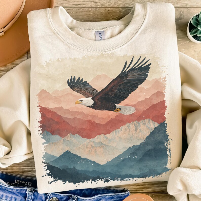 Op de afbeelding: Cr&egrave;mekleurige sweatshirt met een majestueuze Amerikaanse zeearend in vlucht boven een bergketen. De adelaar heeft bruine en witte veren, met een gele snavel. De bergen zijn afgebeeld in tinten rood, blauw en wit.