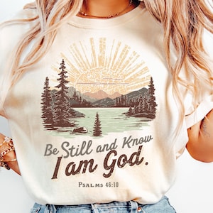 May include: Cream-colored t-shirt featuring a scenic landscape design with a lake, mountains, and trees under a radiant sun. The text "Be Still and Know I am God" is printed below the image, with "PSALMS 46:10" underneath.
