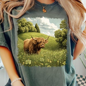 May include: A dark teal t-shirt featuring a detailed illustration of a Highland cow in a grassy field with trees and a cloudy sky. The cow is brown with long hair and curved horns. The scene includes four-leaf clovers.