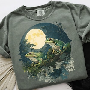 May include: A sage green t-shirt featuring a circular graphic of two green frogs perched on a branch, with a full moon in the background. The design includes forest elements and a dark blue night sky. The shirt has a crew neck and short sleeves.