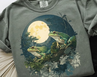 May include: A sage green t-shirt featuring a circular graphic of two green frogs perched on a branch, with a full moon in the background. The design includes forest elements and a dark blue night sky. The shirt has a crew neck and short sleeves.