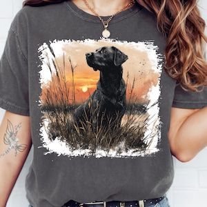 May include: Dark grey t-shirt featuring a black Labrador Retriever illustration. The dog is set against a sunset backdrop with tall grass. The graphic is framed with a white border, creating a striking visual.