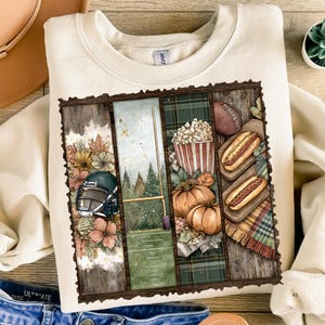 May include: Cream-colored sweatshirt with a football-themed graphic. The graphic features a football helmet, a football field, popcorn, pumpkins, hot dogs, and a football. The design is framed with a wooden border.