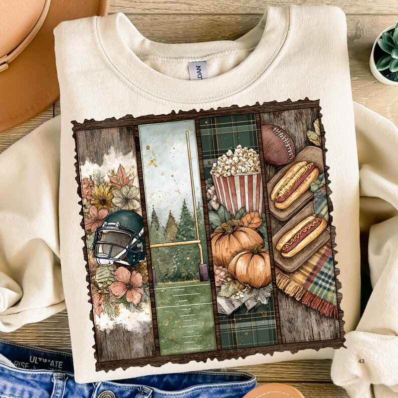 May include: Cream-colored sweatshirt with a football-themed graphic. The graphic features a football helmet, a football field, popcorn, pumpkins, hot dogs, and a football. The design is framed with a wooden border.