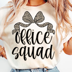 May include: Cream-colored t-shirt featuring a leopard print bow and the words "office squad" in black script. The bow is centered above the text, adding a decorative element to the design. The shirt is a casual wear item.