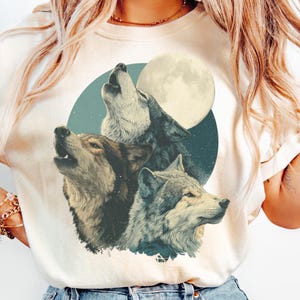 May include: Cream-colored t-shirt featuring a graphic of three wolves howling at a full moon. The wolves are depicted in shades of gray and brown against a teal and starry background. The moon is a bright yellow circle.