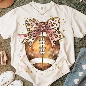 May include: White t-shirt with a football design and a pink leopard print bow. The football is brown with white stripes. Gold and pink accents are scattered around the design. A necklace is visible at the neckline.