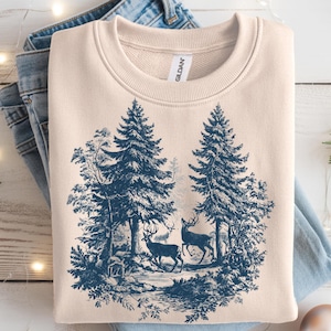 May include: A cream-colored sweatshirt with a blue forest scene featuring two deer and tall pine trees. The design is reminiscent of a vintage print. The sweatshirt is folded on top of a pair of blue jeans.