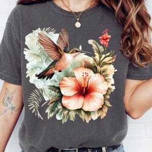 May include: Dark gray t-shirt featuring a colorful hummingbird in flight, set against a backdrop of tropical flowers and foliage. The design includes a large peach hibiscus and various green leaves, creating a vibrant, nature-inspired graphic.