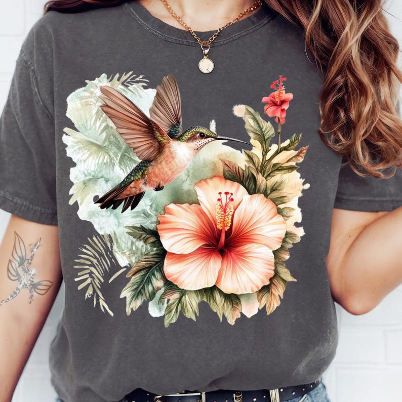 May include: Dark gray t-shirt featuring a colorful hummingbird in flight, set against a backdrop of tropical flowers and foliage. The design includes a large peach hibiscus and various green leaves, creating a vibrant, nature-inspired graphic.