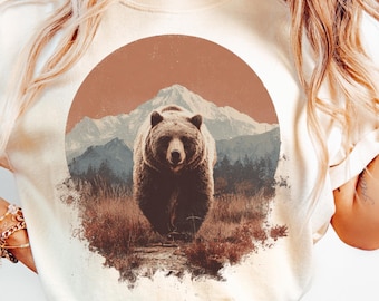 May include: Cream-colored sweatshirt featuring a brown bear illustration set against a mountain landscape within a circular design. The bear is the focal point, with a detailed depiction of its fur and expression. The background includes a mountain range and forest.