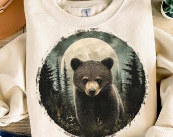 May include: Cream-colored sweatshirt featuring a detailed illustration of a bear cub set against a forest and full moon backdrop. The bear is dark brown with a light brown snout. The design is framed by a circular border.