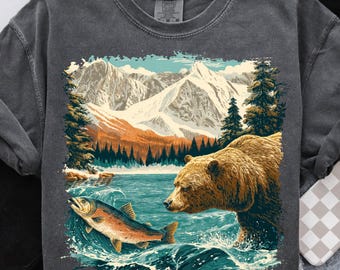 May include: Dark gray sweatshirt featuring a nature scene with a bear and a fish in a lake, with mountains in the background. The bear is brown, the fish is orange, and the water is blue. The sweatshirt has a crew neck.