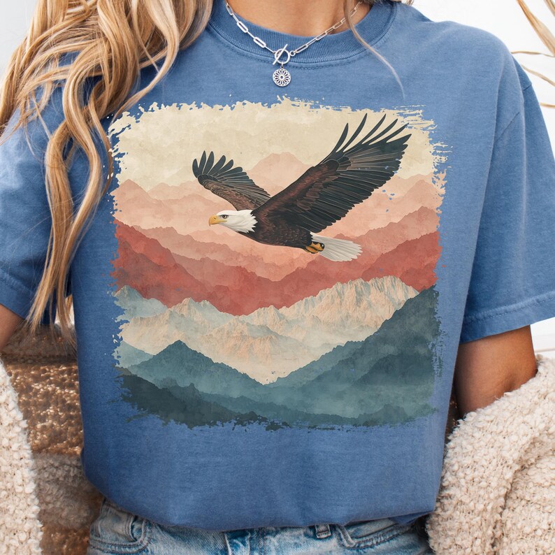 Op de afbeelding: Blauw T-shirt met een afbeelding van een Amerikaanse zeearend die over een bergketen vliegt. De adelaar is bruin en wit. De bergen zijn in tinten rood, bruin en blauw.