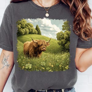 May include: Dark grey t-shirt featuring a detailed illustration of a Highland cow in a grassy field with a blue sky background. The cow is brown with long fur and curved horns. The field has green grass and yellow flowers.