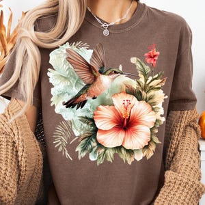 May include: Brown t-shirt featuring a vibrant hummingbird with outstretched wings, set against a backdrop of tropical flowers and foliage. The design includes a large, coral-colored hibiscus and other floral elements.