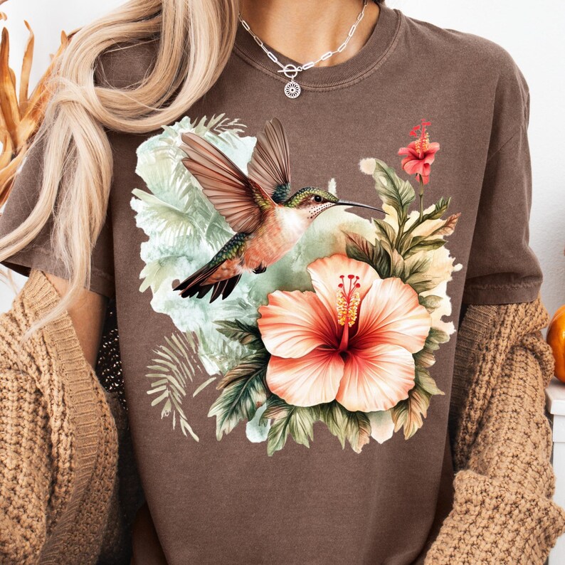 May include: Brown t-shirt featuring a vibrant hummingbird with outstretched wings, set against a backdrop of tropical flowers and foliage. The design includes a large, coral-colored hibiscus and other floral elements.