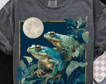 May include: Dark gray sweatshirt featuring a square graphic with two frogs in shades of blue and green, surrounded by foliage, with a full moon in the background. The graphic is framed in gold. The sweatshirt is a Comfort Colors brand.