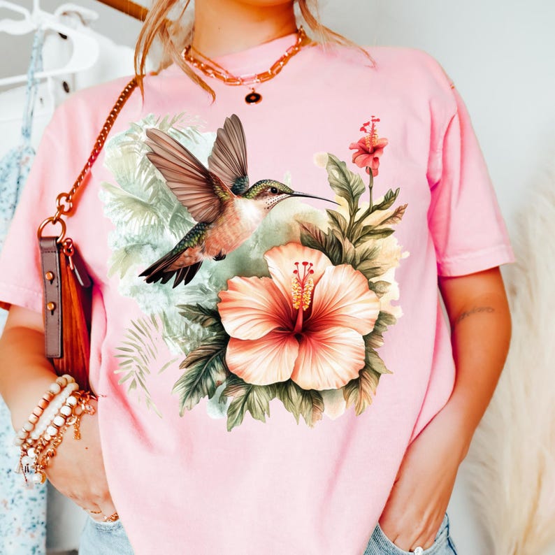 May include: Pink t-shirt featuring a vibrant hummingbird design. The graphic includes a hummingbird with outstretched wings, a large peach-colored hibiscus flower, and lush green foliage. The shirt is a soft, casual style.