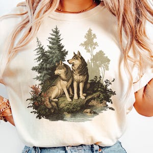 May include: Cream-colored t-shirt featuring a detailed illustration of two wolves in a forest setting. The artwork depicts the wolves near a body of water, surrounded by trees and foliage. The design has a vintage, nature-inspired aesthetic.