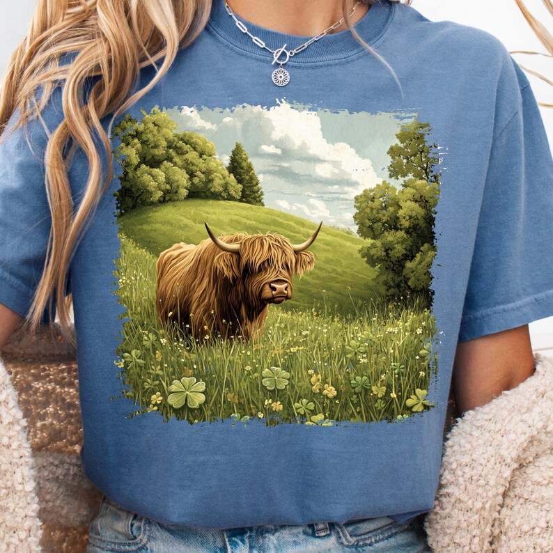 May include: Blue t-shirt featuring a graphic of a Highland cow in a field. The cow is brown with long hair and curved horns. The background shows a green hill, trees, and a cloudy sky. The shirt is made of a soft material.