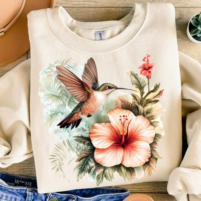 May include: Cream-colored sweatshirt featuring a hummingbird in flight, with a large hibiscus flower and tropical foliage. The hummingbird has brown wings, a green head, and a long beak. The hibiscus is peach and red.
