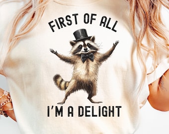 May include: Cream-colored t-shirt featuring a raccoon wearing a top hat and bow tie, with the text "FIRST OF ALL I'M A DELIGHT" in black. The raccoon is depicted with its arms raised. A fun, whimsical design.