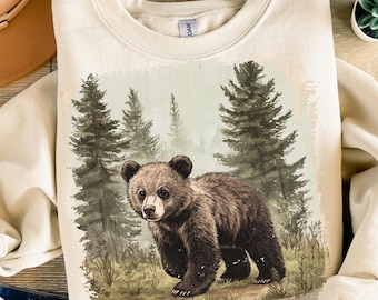 May include: Cream-colored sweatshirt featuring a watercolor illustration of a brown bear in a forest setting. The bear is the focal point, with detailed fur and a curious expression. The background includes pine trees and a misty atmosphere.