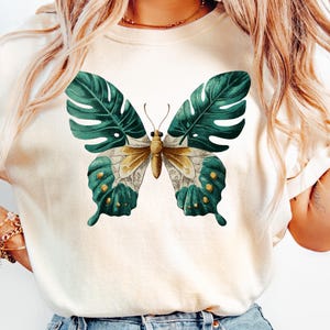 May include: Cream-colored t-shirt featuring a butterfly design. The butterfly has green leaf-like wings with yellow and teal accents. The butterfly's body is a light brown color. The shirt is a casual, everyday wear item.