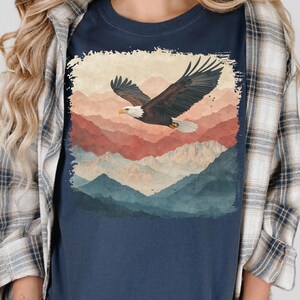 Op de afbeelding: Marineblauwe t-shirt met een afbeelding van een Amerikaanse zeearend in vlucht boven een bergketen. De adelaar heeft bruine en witte veren, met een gele snavel. De bergen zijn afgebeeld in tinten rood, oranje en blauw.