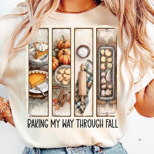 May include: Cream-colored t-shirt featuring a watercolor design of fall baking elements. The design includes pumpkin pie, pumpkins, cookies, a rolling pin, and the text "Baking My Way Through Fall."