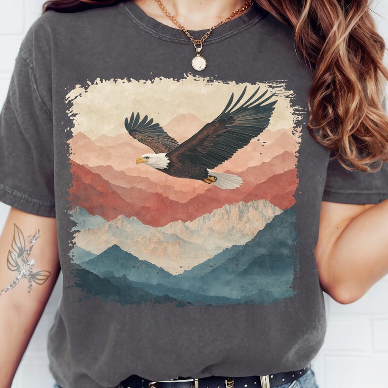 Op de afbeelding: Donkergrijs t-shirt met een afbeelding van een Amerikaanse zeearend die over een bergketen vliegt. De arend heeft bruine en witte veren, met een gele snavel. De bergen zijn in tinten rood, blauw en beige.