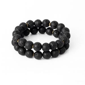 May include: Two black lava stone bead bracelets. Each bracelet is made of round, porous black beads. The beads are strung together with gold-colored spacers. The bracelets are displayed on a white background.