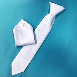 May include: A white tie and pocket square set with embroidered crosses. The tie is long and narrow, and the pocket square is folded neatly. The set is made of a smooth, white fabric and is set against a teal background.