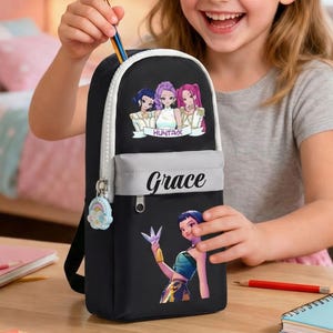 Puede incluir: Mochila negra con parte superior blanca y bolsillo gris, con un gráfico colorido y el nombre "Grace". La mochila tiene cierre de cremallera y un pequeño colgante. Se está colocando un lápiz en la mochila.