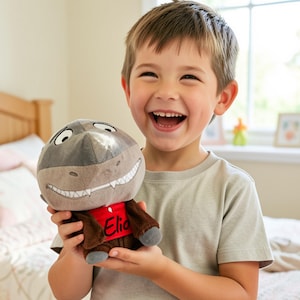Pode incluir: Um brinquedo de pelúcia em forma de tubarão de desenho animado, com cabeça cinza, focinho castanho e uma camisa vermelha com o nome "Elia". O brinquedo é segurado por uma criança sorridente em uma camiseta cinza claro.