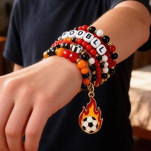 May include: A multi-strand bracelet with red, black, orange, and white beads spelling "FOOTBALL". A gold charm with a soccer ball in flames hangs from the bracelet. The bracelet is worn on a wrist.