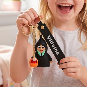 May include: A black keychain with a cartoon villain figure and the word "Villains" on the strap. The figure is holding a small orange and red object. The keychain is attached to a gold-colored ring.