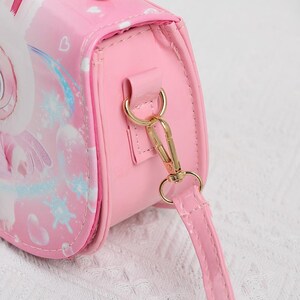 May include: Pink crossbody bag with a cartoon design. The bag features a gold-colored clasp and strap attachments. The strap is pink and the bag has a glossy finish. The design on the bag includes a unicorn and snowflakes.