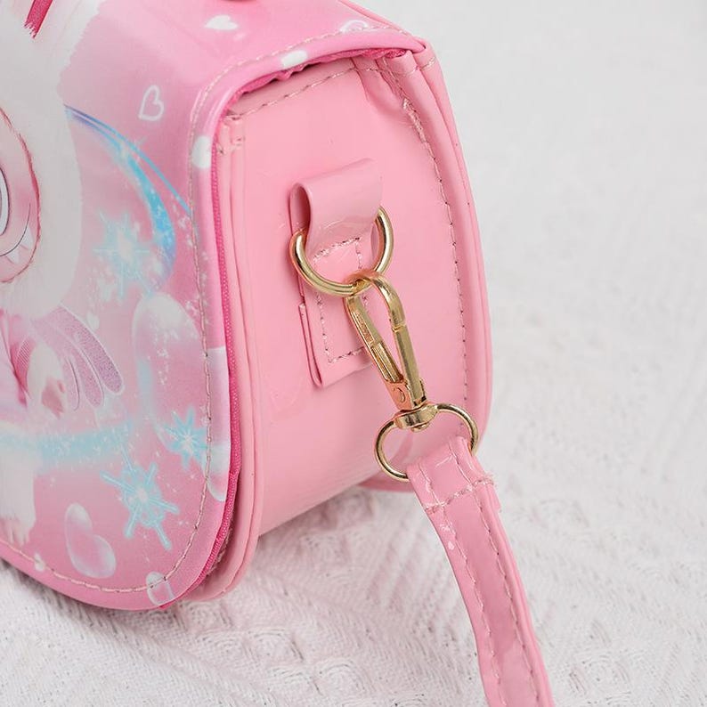 May include: Pink crossbody bag with a cartoon design. The bag features a gold-colored clasp and strap attachments. The strap is pink and the bag has a glossy finish. The design on the bag includes a unicorn and snowflakes.