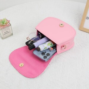 May include: A pink, open, small purse with a gold clasp, revealing contents including a smartphone, glasses, and beauty products. The purse is on a light-colored surface. A small succulent plant in a glass container is visible in the background.
