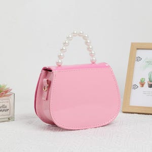 May include: A glossy pink handbag with a curved shape and a handle made of white faux pearls. The bag has a flap closure and gold-colored hardware. The bag is sitting on a white surface.