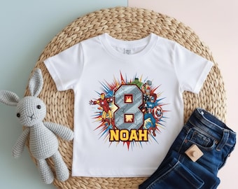 May include: White t-shirt featuring a colorful graphic of the number 8 with superhero characters and the name "NOAH" in yellow. The design includes Iron Man, Captain America, and the Hulk. The shirt is paired with blue denim jeans.