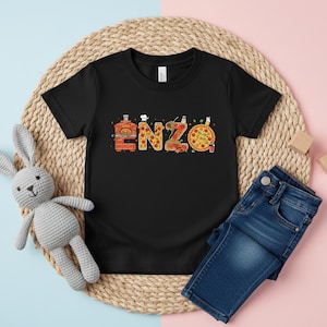 May include: Black t-shirt with the name "ENZO" in a pizza-themed design. The letters are styled with pizza oven, pizza slices, and a whole pizza. The shirt is paired with blue denim jeans and a stuffed bunny.
