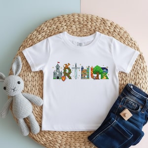 May include: White t-shirt with the name "ARTHUR" printed across the front in colorful, whimsical illustrations. The letters are designed with images of a castle, dragon, sword, and tree. The shirt is paired with blue jeans.