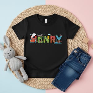 May include: Black short-sleeved t-shirt with the name "HENRY" in colorful, playful letters. The letters are designed with train, house, and road motifs. A pair of blue denim jeans and a gray stuffed bunny are also in the image.