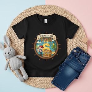 May include: Black t-shirt with a pirate-themed graphic for a third birthday. The design includes a pirate, treasure chest, and the name "Finn" with the number "3". Paired with blue denim jeans.