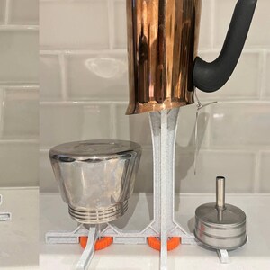 May include: A collection of three kitchen utensils: a copper kettle, a stainless steel container, and a small stainless steel container with a lid. They are supported by white and orange 3D-printed stands.